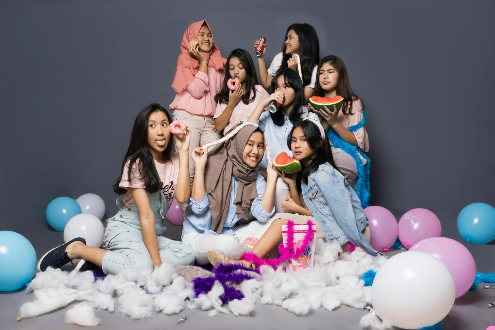 Group of happy girls taking during a photoshoot among colorful balloons while eating snacks 